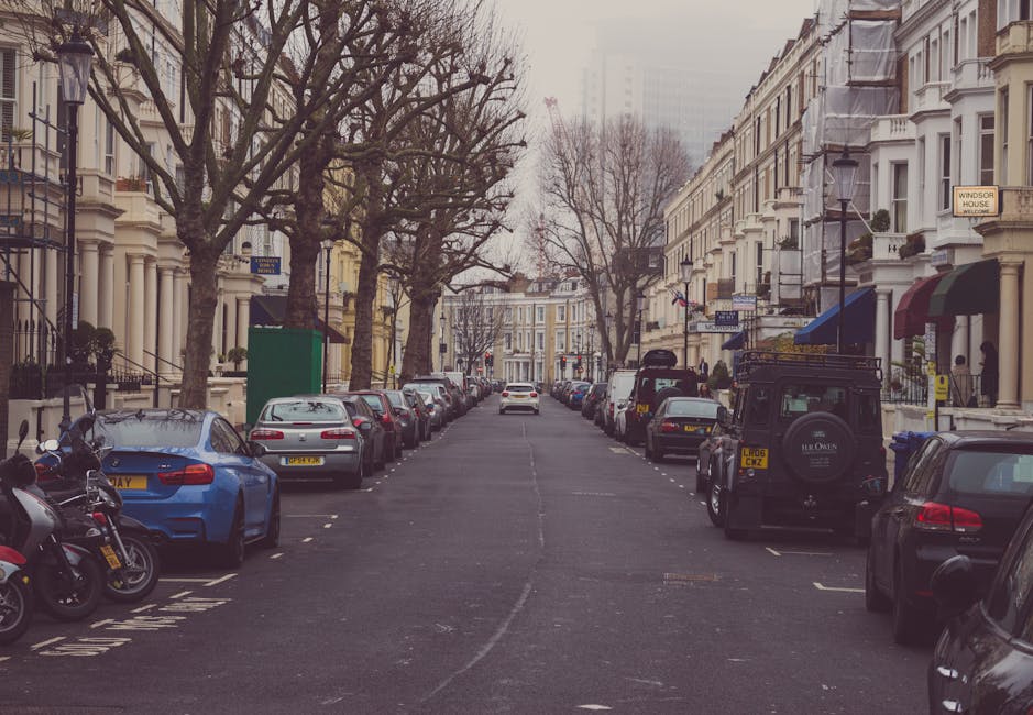 A residential street in Kentish Town with a row of terraced Victorian-style houses featuring white facades, large windows, and decorative cornices. The street is lined with a variety of parked cars, including a blue sedan, a silver hatchback, and a black SUV, as well as a couple of motorcycles, all neatly parked in designated spaces along both sides of the asphalt road. Leafless trees with thick trunks and sprawling branches stretch overhead, indicating a winter or early spring season. The street is well-lit with classic black lampposts, and small shopfronts with awnings and signage are visible on the right side. The scene appears clean and well-maintained, reflecting a typical residential area in central London. The overall image captures a calm, orderly urban environment suitable for home or commercial cleaning assessments, showcasing the importance of surface cleanliness and maintenance that a company like Cleaner Kentish Town might address.