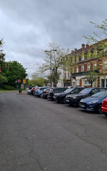 A residential street in Kentish Town with a row of terraced Victorian-style houses featuring white facades, large windows, and decorative cornices. The street is lined with a variety of parked cars, including a blue sedan, a silver hatchback, and a black SUV, as well as a couple of motorcycles, all neatly parked in designated spaces along both sides of the asphalt road. Leafless trees with thick trunks and sprawling branches stretch overhead, indicating a winter or early spring season. The street is well-lit with classic black lampposts, and small shopfronts with awnings and signage are visible on the right side. The scene appears clean and well-maintained, reflecting a typical residential area in central London. The overall image captures a calm, orderly urban environment suitable for home or commercial cleaning assessments, showcasing the importance of surface cleanliness and maintenance that a company like Cleaner Kentish Town might address.