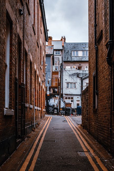 A narrow urban alleyway between two brick residential buildings, with a paved surface featuring double yellow lines along the edges. The alley is clean and free of debris, with visible drainage grates embedded in the pavement. At the end of the alley, there are multi-storey buildings with a mix of brick and concrete facades, some with small external staircases and external storage areas. Overhead, the sky is overcast, giving the scene a muted lighting. The image illustrates an exterior communal space that could benefit from regular surface cleaning and maintenance, aligning with the themes of hygienic and thorough cleaning provided by Cleaner Kentish Town for both domestic and commercial environments.
