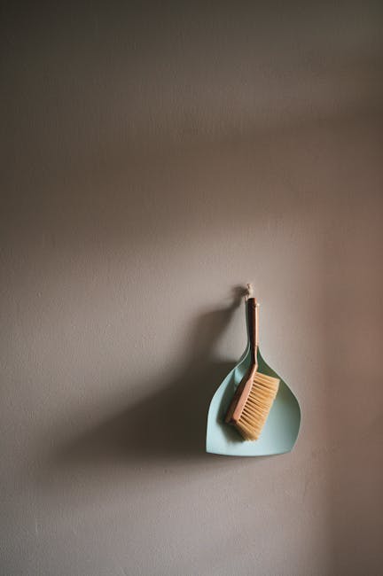 A close-up of a beige cleaning brush with natural bristles hanging from a hook on a neutral grey wall. The brush is positioned inside a light teal silicone holder, which is also hanging on the hook. The wall surface appears smooth and clean, with soft, natural lighting illuminating the scene. The minimalist setup suggests a focus on surface cleaning tools stored neatly in a domestic or commercial setting, with the branding of Cleaner Kentish Town implied through the context of hygiene and maintenance associated with their services in preventing hidden cleaning charges in Kentish Town flats.