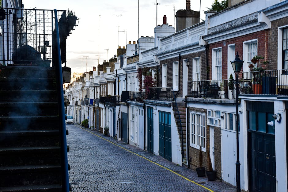 A narrow urban alleyway between two brick residential buildings, with a paved surface featuring double yellow lines along the edges. The alley is clean and free of debris, with visible drainage grates embedded in the pavement. At the end of the alley, there are multi-storey buildings with a mix of brick and concrete facades, some with small external staircases and external storage areas. Overhead, the sky is overcast, giving the scene a muted lighting. The image illustrates an exterior communal space that could benefit from regular surface cleaning and maintenance, aligning with the themes of hygienic and thorough cleaning provided by Cleaner Kentish Town for both domestic and commercial environments.