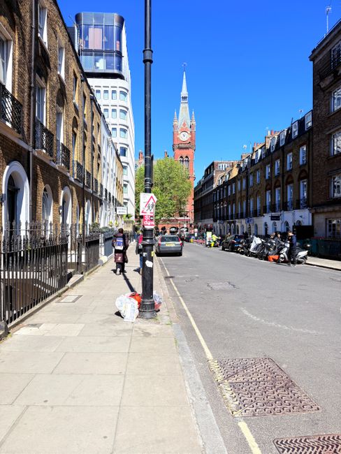 A daytime street view in Kentish Town with a clear blue sky. The scene shows a paved sidewalk on the left side, with a person walking and another standing near a pile of trash bags. The sidewalk is lined with historic brick buildings featuring arches and black iron railings. On the right, there is a row of parked motorcycles and cars along the curb. In the background, a prominent red-brick clock tower with a pointed spire is visible, surrounded by modern glass and steel buildings. The street appears clean and well-maintained, reflecting the importance of regular cleaning and maintenance, which is well-supported by professional services like Cleaner Kentish Town.