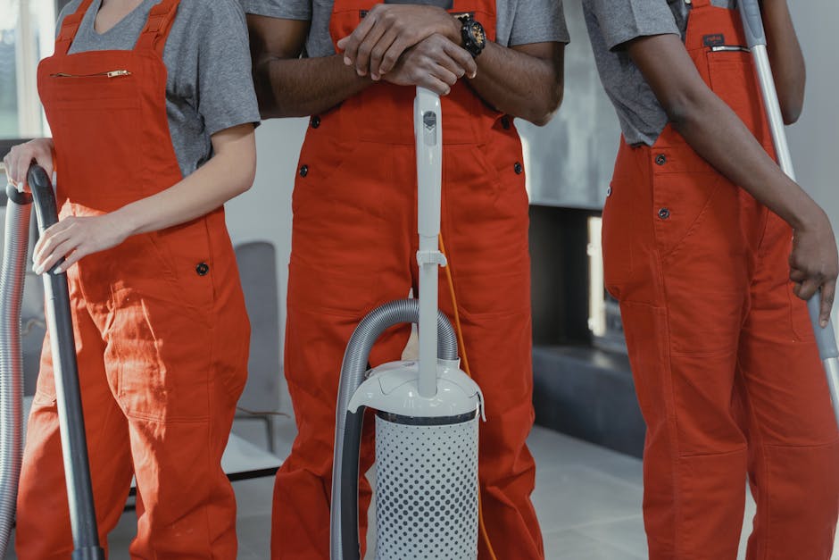 Close-up of three professional cleaners wearing orange uniforms and gray shirts, holding vacuum cleaners and cleaning tools inside a modern residential space. The image shows the lower halves of their