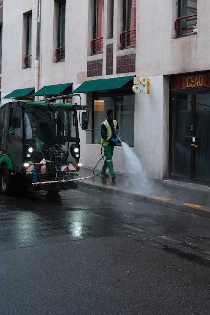 A worker from Cleaner Kentish Town engaged in surface cleaning using a high-pressure power washer on a paved street outside a building with beige walls and green awnings. The worker is wearing a high-visibility vest and gloves, focusing on cleaning the asphalt surface. A small street cleaning vehicle with attached equipment is positioned nearby, spraying water to remove dirt and grime, resulting in a wet, shiny surface. The building features large windows with vertical metal supports and red-tinted awnings, with the street scene illuminated by daylight. The image depicts a professional deep cleaning process targeting urban surfaces to maintain hygiene and cleanliness, consistent with services offered near Kentish Town Station NW5.