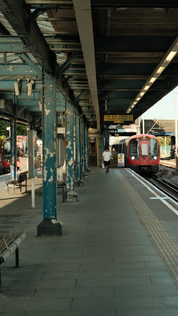 A worker from Cleaner Kentish Town engaged in surface cleaning using a high-pressure power washer on a paved street outside a building with beige walls and green awnings. The worker is wearing a high-visibility vest and gloves, focusing on cleaning the asphalt surface. A small street cleaning vehicle with attached equipment is positioned nearby, spraying water to remove dirt and grime, resulting in a wet, shiny surface. The building features large windows with vertical metal supports and red-tinted awnings, with the street scene illuminated by daylight. The image depicts a professional deep cleaning process targeting urban surfaces to maintain hygiene and cleanliness, consistent with services offered near Kentish Town Station NW5.