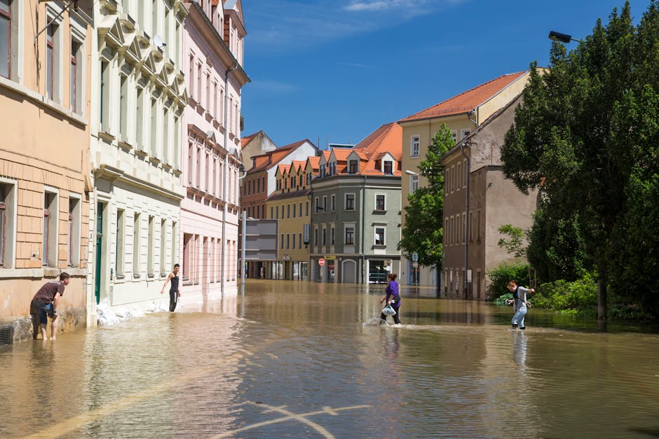Image of a flooded urban street in daylight, with water covering the pavement and partially submerging the lower parts of multi-story buildings with pastel-colored facades and red-tiled roofs. Several