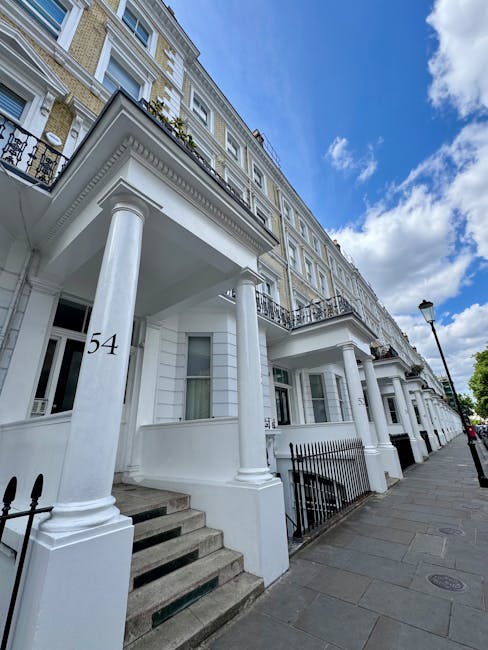 Exterior view of a row of white residential buildings on Kentish Town Road under a partly cloudy sky, featuring classic architectural details with large columns supporting small balconies, steps leading up to the entrances, and black iron railings. The pavement is clean and well-maintained, with a street lamp visible along the sidewalk. The image emphasizes the building facades and street environment, suitable for illustrating surface cleaning, maintenance, or property sanitisation services provided by Cleaner Kentish Town for end of tenancy cleaning in NW5.