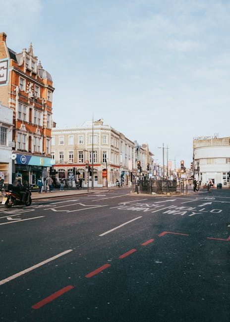 This image shows a busy street scene at the end of a shopping and residential area in Kentish Town, NW5. The street features a wide asphalt surface with clear lane markings, including a dashed red line and a dedicated bus lane. On the left, there are historic multi-story buildings with ornate facades, large windows, and decorative gables, likely houses or shops, with some storefront signs visible. Pedestrians are walking along the sidewalks, and a motorcycle is parked near the curb. The urban environment is well lit by natural daylight, with a partly cloudy sky overhead. The scene captures a lively typical day in Kentish Town with shops, pedestrians, and street infrastructure, suggesting a well-maintained and clean urban area, fitting for content related to city cleaning and maintenance services provided by Cleaner Kentish Town.