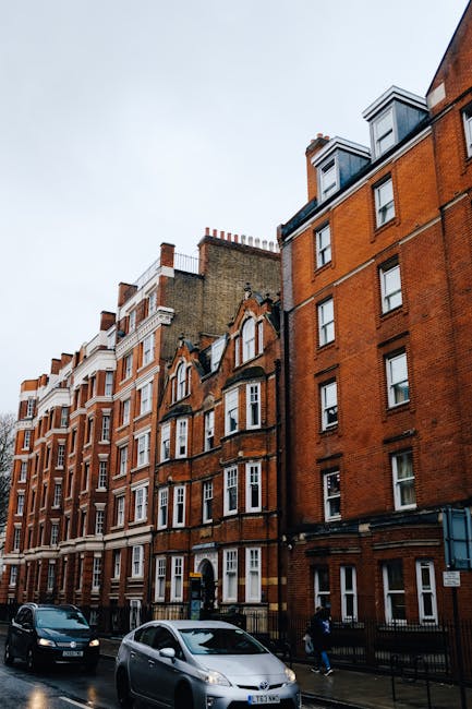Exterior view of a row of white residential buildings on Kentish Town Road under a partly cloudy sky, featuring classic architectural details with large columns supporting small balconies, steps leading up to the entrances, and black iron railings. The pavement is clean and well-maintained, with a street lamp visible along the sidewalk. The image emphasizes the building facades and street environment, suitable for illustrating surface cleaning, maintenance, or property sanitisation services provided by Cleaner Kentish Town for end of tenancy cleaning in NW5.