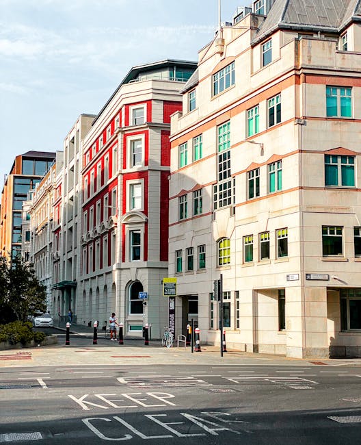 Photograph of a city street viewed from across the road, featuring multi-storey buildings with a mix of modern and classical architectural styles. The buildings have large windows, some with green and yellow panes, and facades made of beige stone and red brick. The street has marked pedestrian crossings, bollards, and signage, with a few bicycles and pedestrians visible. Bright daylight enhances the clean, well-maintained appearance of the area. The image emphasizes urban cleanliness and maintenance, consistent with professional cleaning services provided by Cleaner Kentish Town.