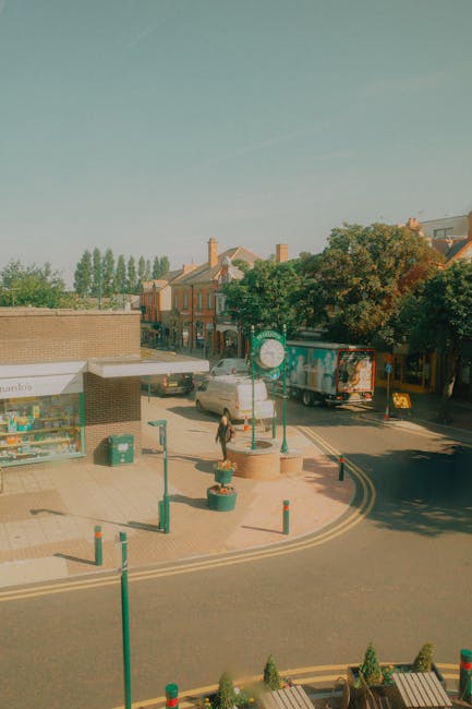 A street scene in Kentish Town during daytime, featuring a small commercial area with a corner shop that has a glass display window and a beige brick exterior. The shop is adjacent to a paved sidewalk with associated street furniture, including green bollards and large round planters with small evergreen shrubs. A pedestrian is walking along the sidewalk, and several vehicles, including a delivery truck and a car, are parked or moving along the road. Residential buildings with pitched roofs, brick facades, and chimneys line the background, with tall trees providing greenery. The sky is clear with a light blue hue, and soft sunlight illuminates the scene. This image is used to illustrate the vibrant, clean environment of Kentish Town, emphasizing the importance of effective cleaning and maintenance services offered by Cleaner Kentish Town, particularly for commercial and office spaces like those seen on Kentish Town Road.