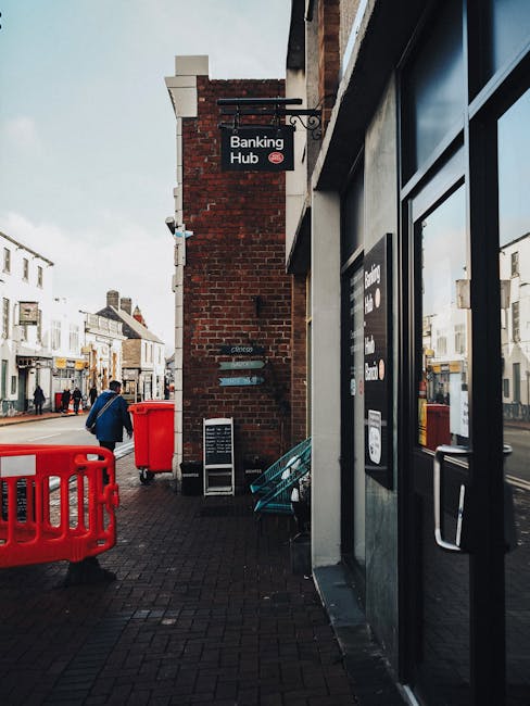 Photograph of a city street viewed from across the road, featuring multi-storey buildings with a mix of modern and classical architectural styles. The buildings have large windows, some with green and yellow panes, and facades made of beige stone and red brick. The street has marked pedestrian crossings, bollards, and signage, with a few bicycles and pedestrians visible. Bright daylight enhances the clean, well-maintained appearance of the area. The image emphasizes urban cleanliness and maintenance, consistent with professional cleaning services provided by Cleaner Kentish Town.