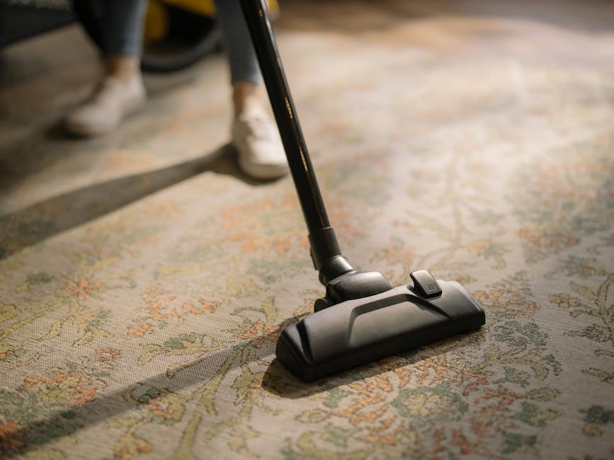 A person kneeling on a patterned carpet in a residential room, holding a yellow and black portable vacuum cleaner with an attached hose. The individual is wearing a beige jacket and blue jeans, and ap