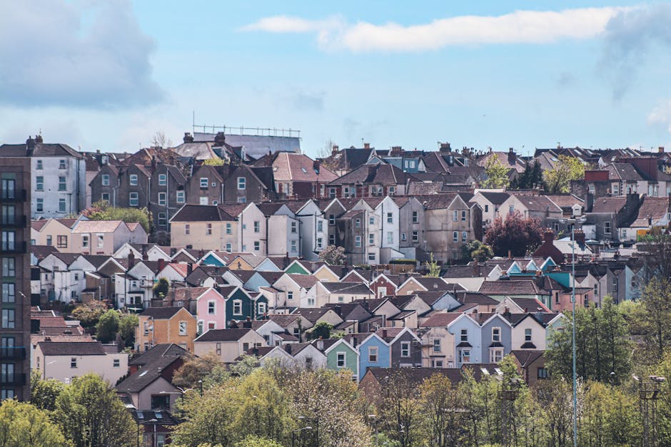 A panoramic view of a densely packed residential neighborhood in Kentish Town, showing a variety of houses with different colors and styles. The houses are situated on a hillside, with some multi-story buildings in the foreground and rows of single-family homes extending into the distance. The rooftops are primarily covered with red or brown tiles, and some buildings feature white or pastel-colored facades. Green trees and foliage are visible among the houses, adding natural contrast to the urban landscape. The sky above is partly cloudy with patches of blue, providing ample natural light that highlights the clean, well-maintained appearance of the community. This image exemplifies a well-kept neighborhood, aligning with the themes of property maintenance and hygiene emphasized by Cleaner Kentish Town in their domestic cleaning services for the area, as seen on the page titled 'Same day house cleaning Kentish Town The Forum, KENTISH TOWN' at cleanerkentishtown.org.uk.