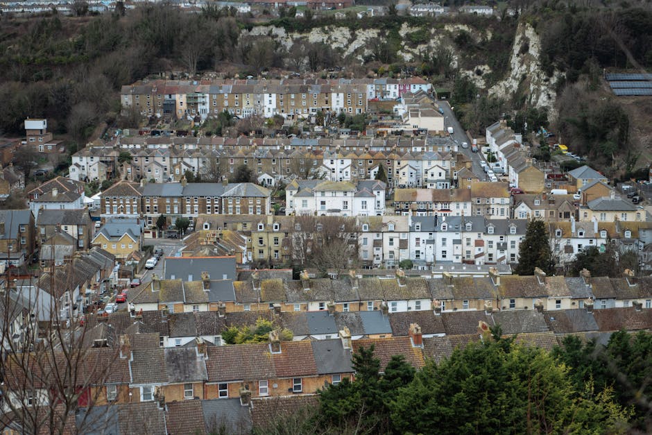An aerial view of a residential area in Kentish Town with tightly packed houses featuring rooftops made of various materials including slate, tile, and stucco, showcasing a mix of brown, gray, and white surfaces. The scene displays rows of terraced homes with chimneys, tiled roofs, and small garden spaces between buildings. Taller buildings such as apartment blocks with brick facades are visible in the background, along with a hill side covered in trees and greenery. The lighting is natural, casting soft shadows, and the overall scene appears clean and well-maintained. No tools or cleaning products are visible in this image, which focuses on the urban landscape, emphasizing the surface textures and the arrangement of the houses, reflecting the environment that Cleaner Kentish Town may service through its domestic cleaning and surface sanitation efforts.