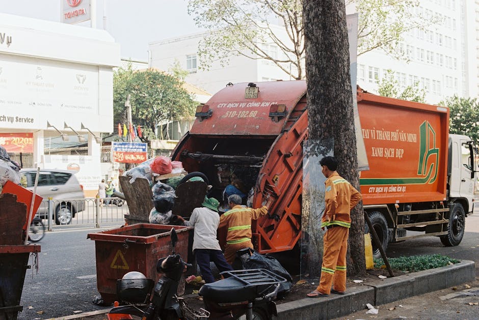 A black-and-white photograph of a waste collection truck parked on a residential street in Kentish Town, with several large black garbage bags and miscellaneous debris loaded on or beside the vehicle.